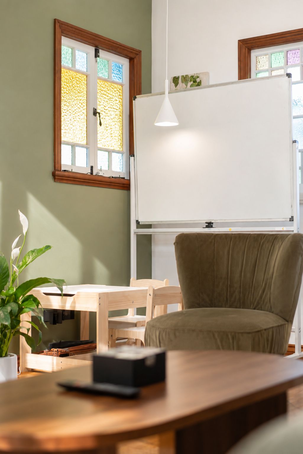 A room with a whiteboard, a wooden desk, a green chair, and a window with stained glass.