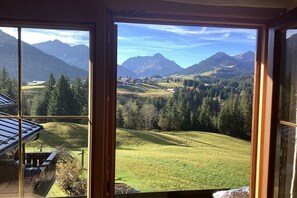 Blick durch ein Fenster auf eine gebirgige Landschaft mit einem Haus und einem Feld.