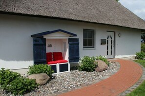 Ein weißes Gebäude mit einem Strohdach, einer kleinen Veranda mit roten Kissen und einem Ziegelweg, der zum Eingang führt.