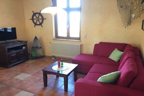 Un salon avec un canapé rouge, une table basse en bois, un téléviseur posé sur un meuble, un décor représentant un gouvernail de bateau et une fenêtre avec des stores.