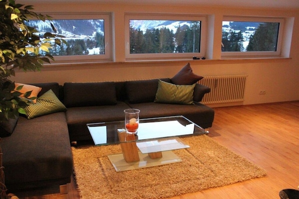 Una sala de estar con un sofá de color oscuro, una mesa de centro de vidrio y vistas a montañas a través de tres ventanas.
