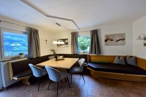 A room with a dining table, chairs, a bench, and a sofa. There are windows with curtains, a cross on the wall, and a painting. - Apartment Steinberger (Ramsau im Zillertal)