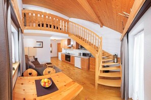 Een houten trap leidt naar een hoger gelegen verdieping, met een eetgedeelte met tafel en stoelen, en een keuken met moderne apparatuur.