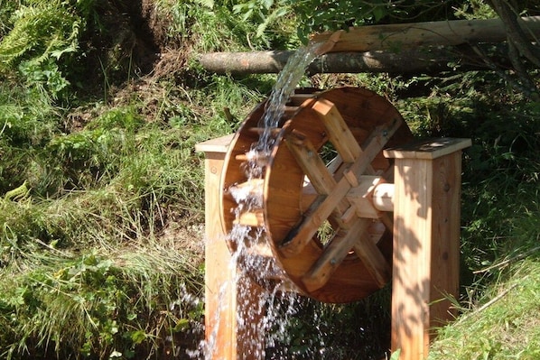 A wooden water wheel with water flowing over it, set in a natural, grassy environment.