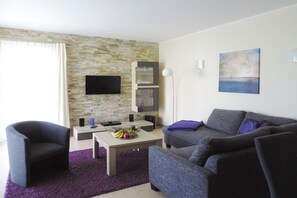A modern living room with a stone wall, a flat-screen TV, a sofa, a coffee table with fruit, and a bookshelf. - Holiday home with 2 bedrooms (Göhren-Lebbin)