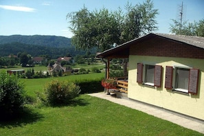 Ein eingeschossiges Gebäude mit einem geneigten Dach, einer Veranda mit einer Bank und Blick auf eine grüne Landschaft mit Bäumen und einer entfernten Hügelkette.