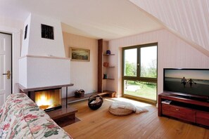 Ein gemütliches Wohnzimmer mit Kamin, Fernseher und großem Fenster mit Blick ins Freie.