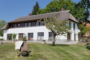 A two-story house with a thatched roof, a white exterior, and a small garden with a tree and a chair. - Holiday apartment with 1 bedroom (Usedom)