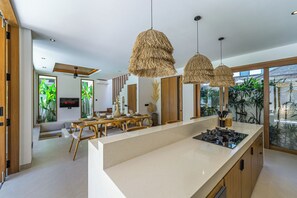 A modern kitchen with a central island, a dining area with a wooden table, and a living space with a sofa and TV. - Villa Paraiso (Umalas)