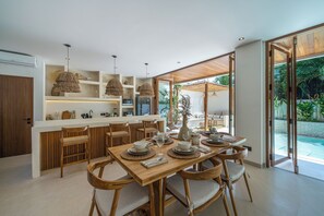 Eine moderne Küche mit Holzschränken, eine Essecke mit Holztisch und -stühlen sowie ein Blick auf ein Schwimmbecken durch große Glastüren.