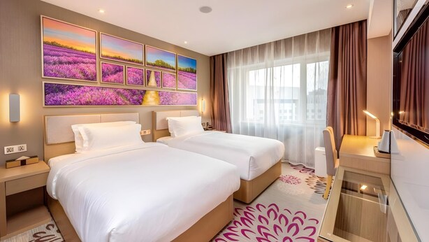 A hotel room with two beds, a large window with curtains, a flat-screen TV, and a wall with multiple framed pictures. - Lavande Hotels Harbin Railway Station Zhanqian Square (Harbin)