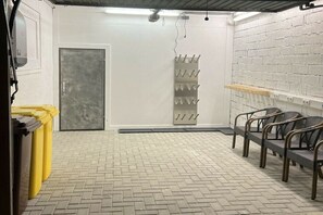Ein Zimmer mit einem Kachelboden, einer Tür, einem Metallgestell mit Haken und einer Reihe von Stühlen.