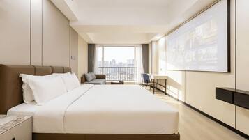 A hotel room with a large bed, a desk, a chair, and a view of the city through the window.