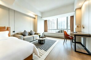 A modern hotel room with a bed, sofa, desk, and large windows offering a city view.