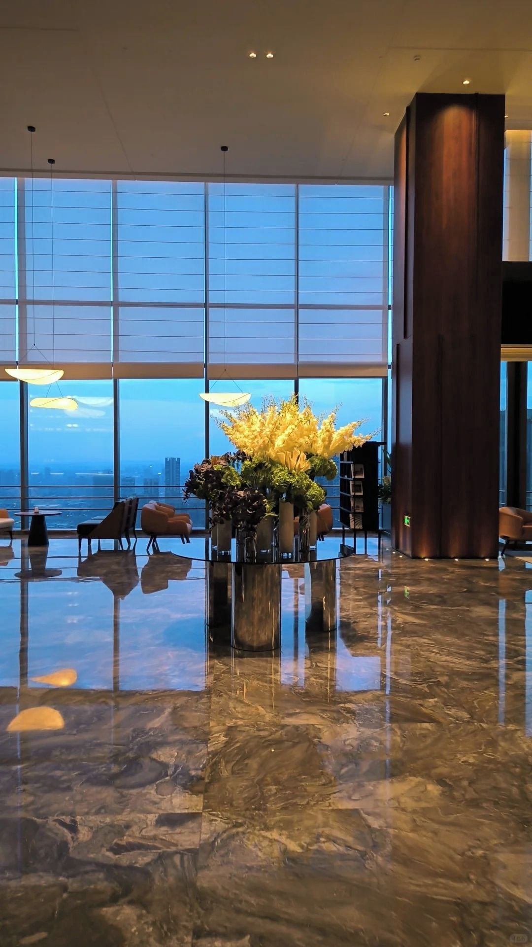 Un hall d’entrée spacieux avec un sol en marbre, de grandes fenêtres donnant sur la ville et un arrangement floral central.