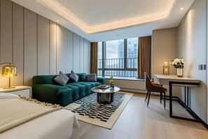 Quarto de hotel moderno com sofá verde, mesa de centro redonda pequena, escrivaninha com cadeira e uma grande janela com cortinas.