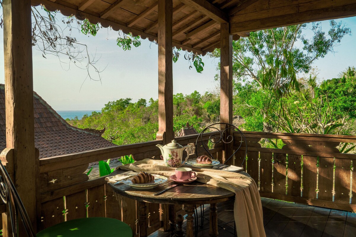 A wooden deck with a table set for breakfast, overlooking a lush landscape and the sea.