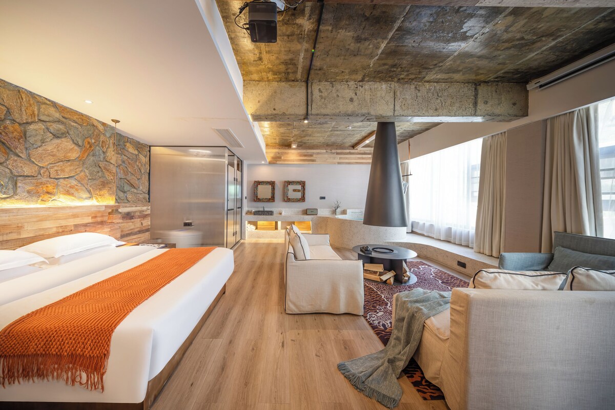 Une chambre d’hôtel moderne avec un mur d’accent en pierre, un grand lit, un coin salon et une vue sur la ville.