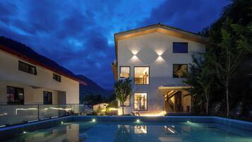 Uma casa moderna de dois andares com piscina, grandes janelas e vista para as montanhas.