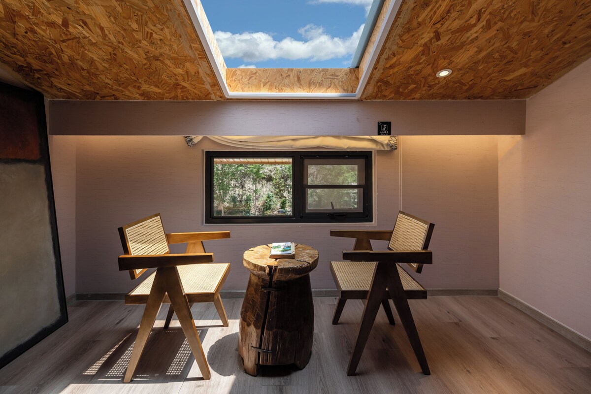 A room with a skylight, two wooden chairs, a round wooden table, and a window with a view of trees.