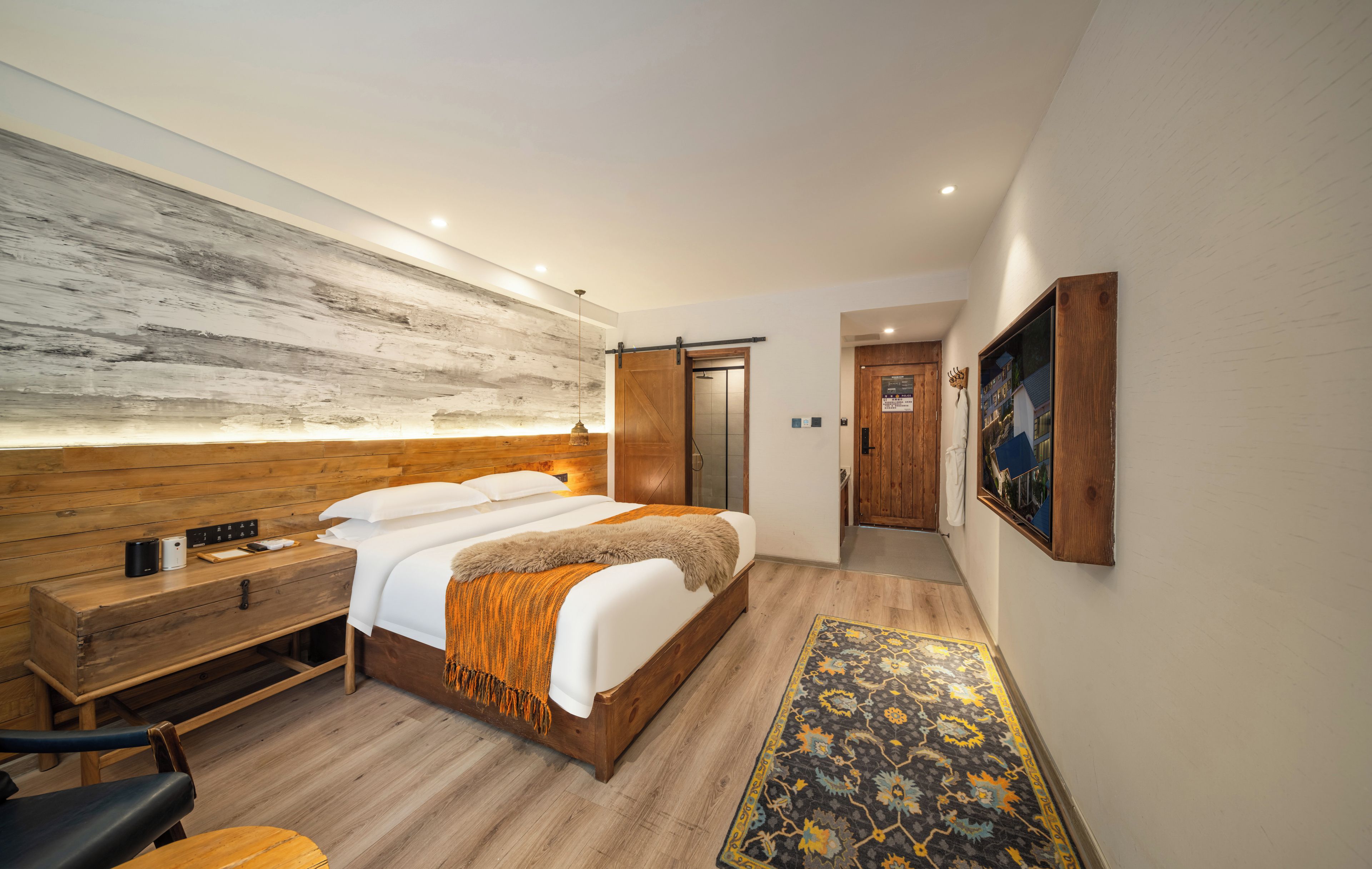 Habitació d'hotel moderna amb un capçal de llit de fusta, un llit gran amb roba de llit blanca, una taula de nit amb una làmpada, una banca de fusta i una catifa amb dibuixos.