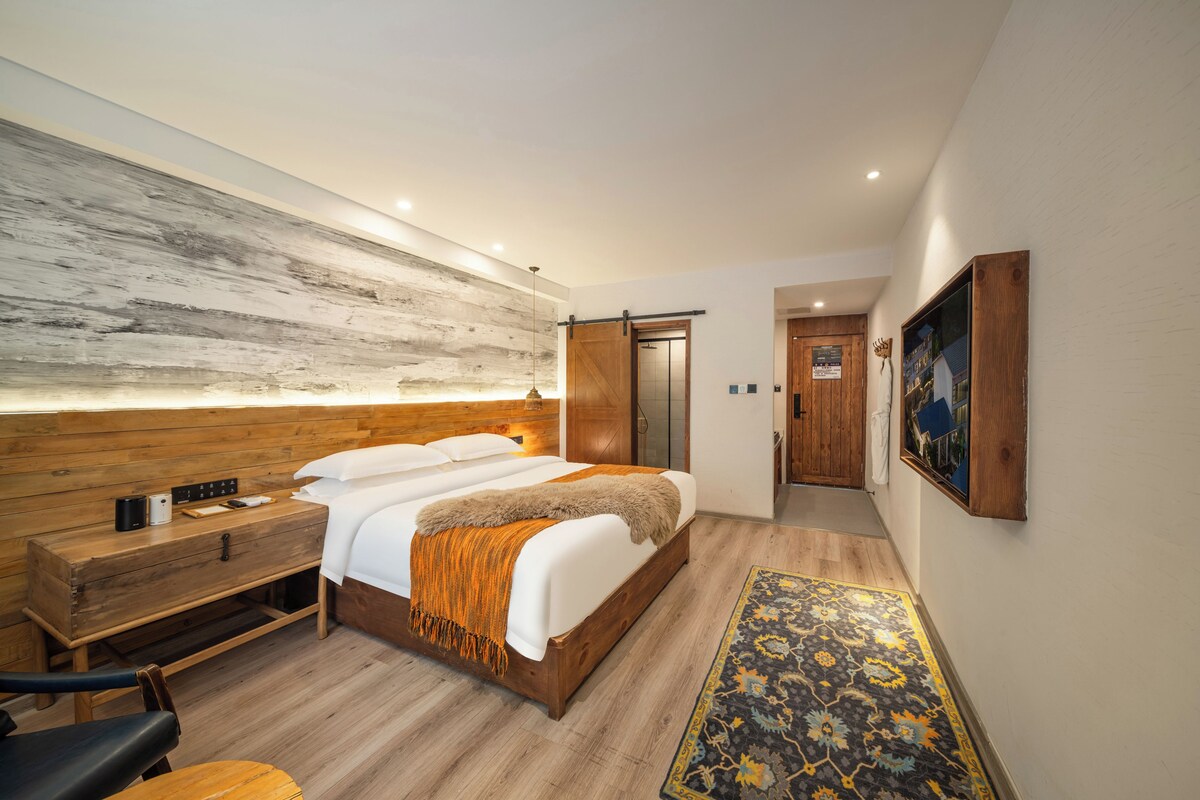 A modern hotel room with a wooden headboard, a large bed with white linens, a bedside table with a lamp, a wooden bench, and a patterned rug.