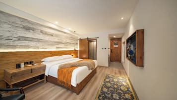 Habitació d'hotel moderna amb un capçal de llit de fusta, un llit gran amb roba de llit blanca, una taula de nit amb una làmpada, una banca de fusta i una catifa amb dibuixos.