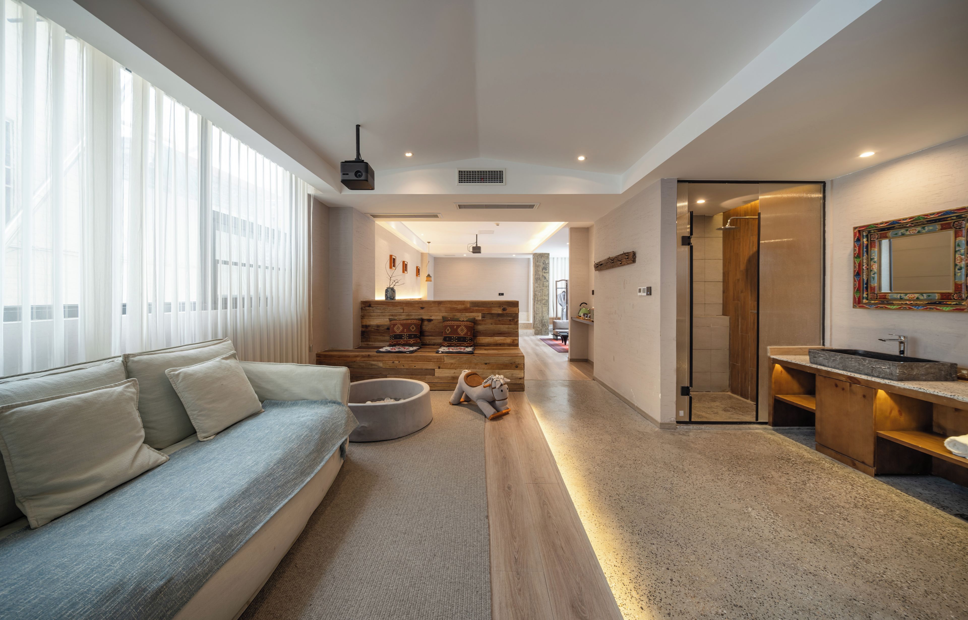 A modern living room with a sofa, a pet bed, and a wooden bench. There is a hallway leading to a bathroom with a shower and a sink.