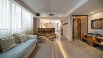 A modern living room with a sofa, a pet bed, and a wooden bench. There is a hallway leading to a bathroom with a shower and a sink.