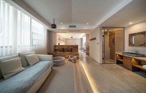 A modern living room with a sofa, a pet bed, and a wooden bench. There is a hallway leading to a bathroom with a shower and a sink.