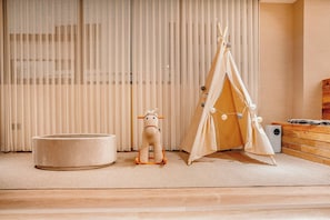A room with a large window, a wooden floor, a round bed, a small wooden horse toy, and a white teepee.