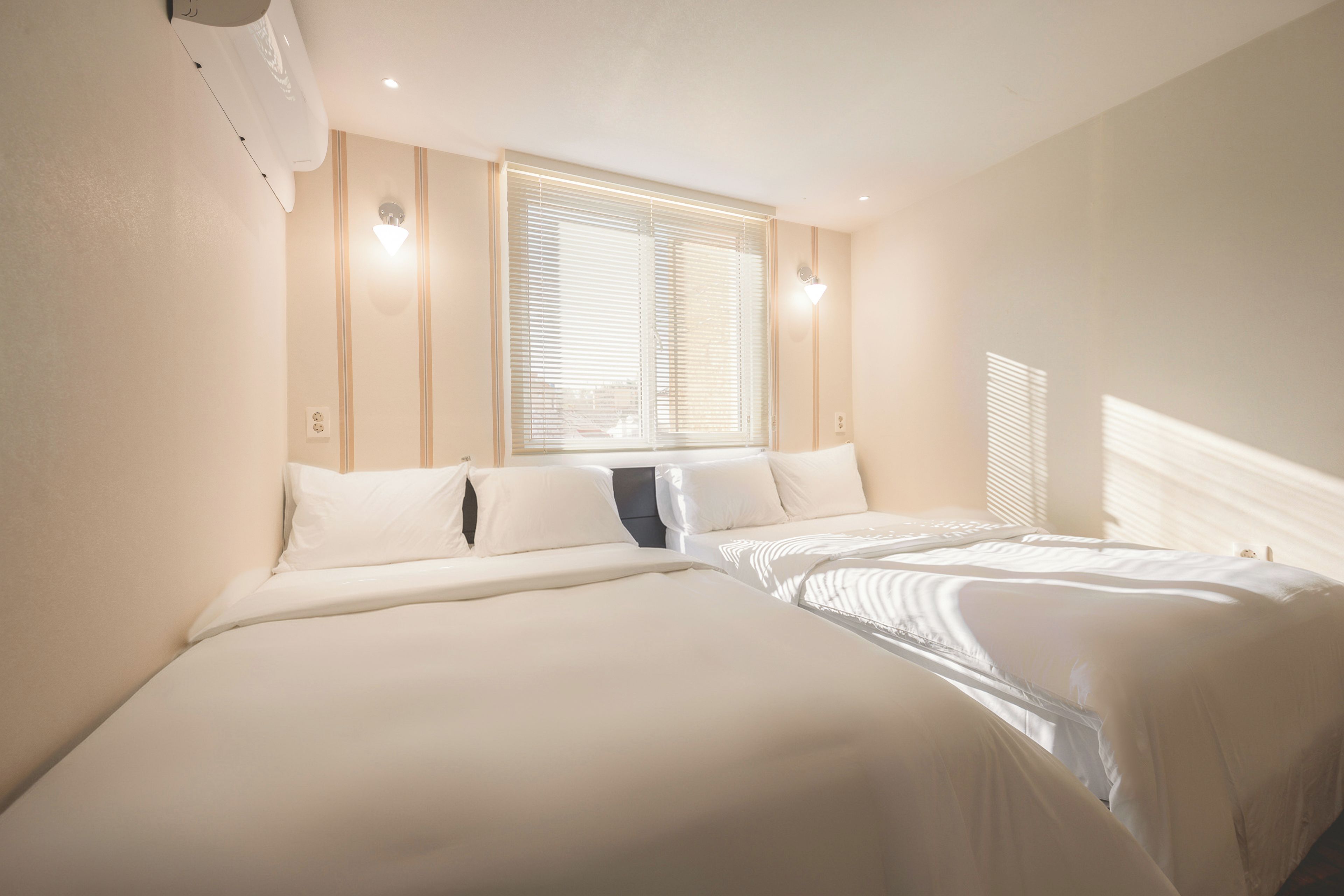 A hotel room with two beds, a window with blinds, and a wall-mounted light fixture.