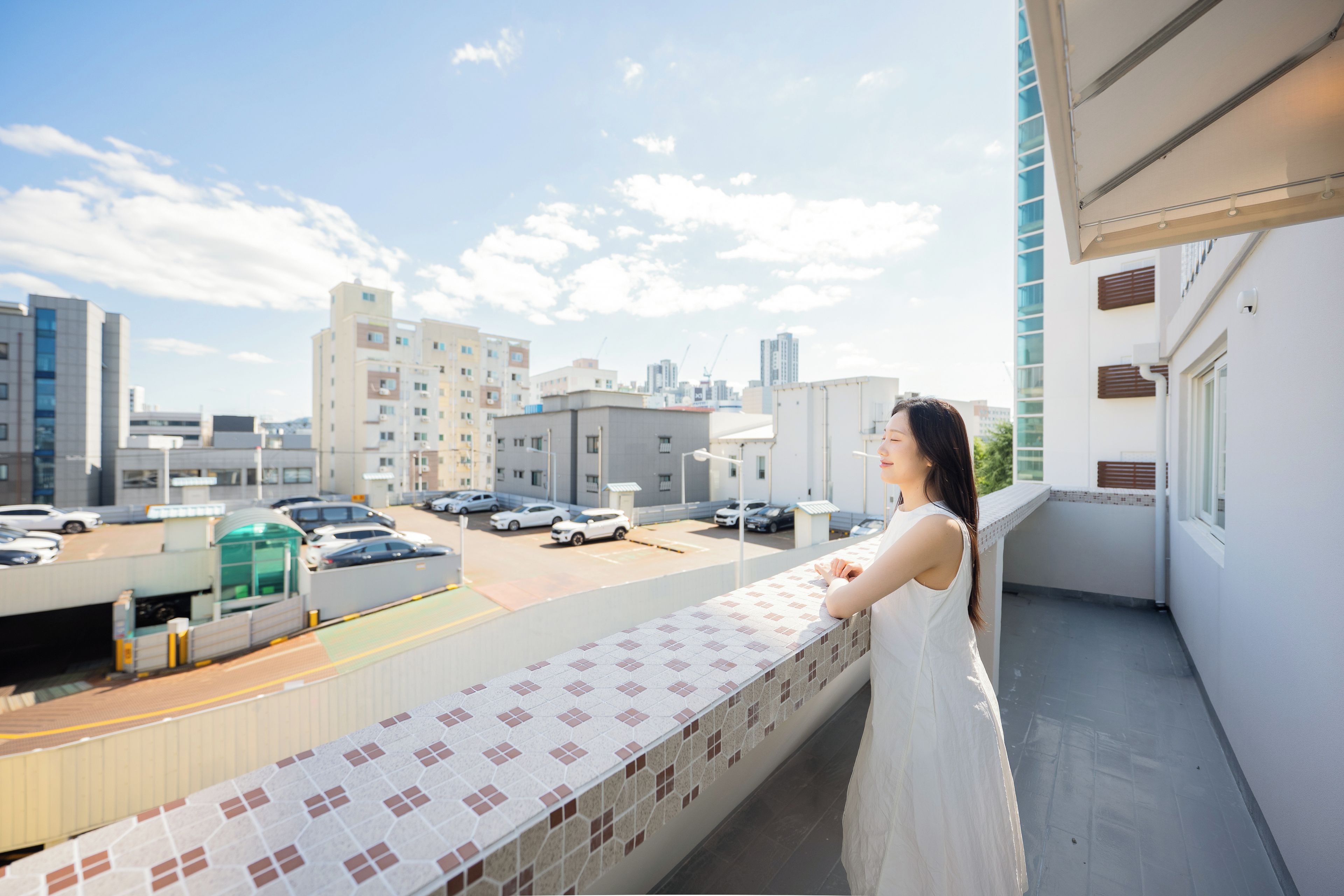 A balcony with a tiled floor and a view of a parking lot and buildings.