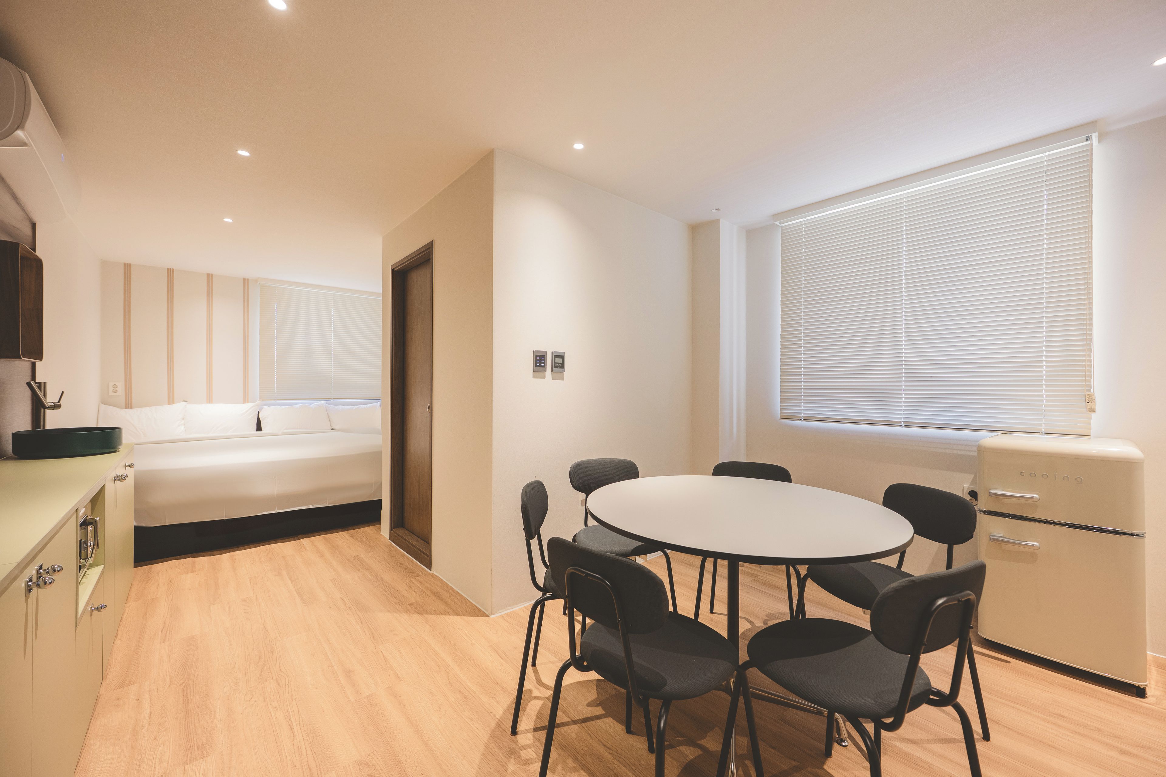 Una habitación de hotel moderna con cama, una mesa de comedor con sillas, kitchenette y un gran ventanal con persianas.