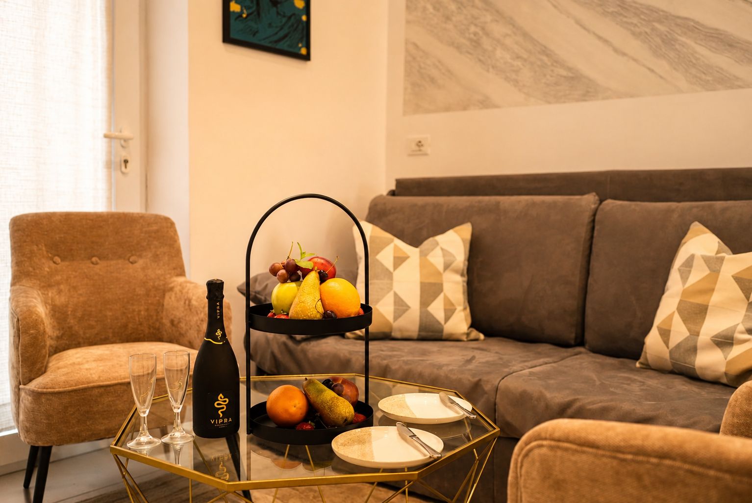 Una sala de estar con un sillón marrón, un sofá con cojines estampados, una mesa de centro de vidrio con una botella y vasos, y una cesta de frutas.