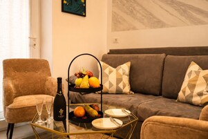 A living room with a brown armchair, a sofa with patterned cushions, a glass coffee table with a bottle and glasses, and a fruit basket.