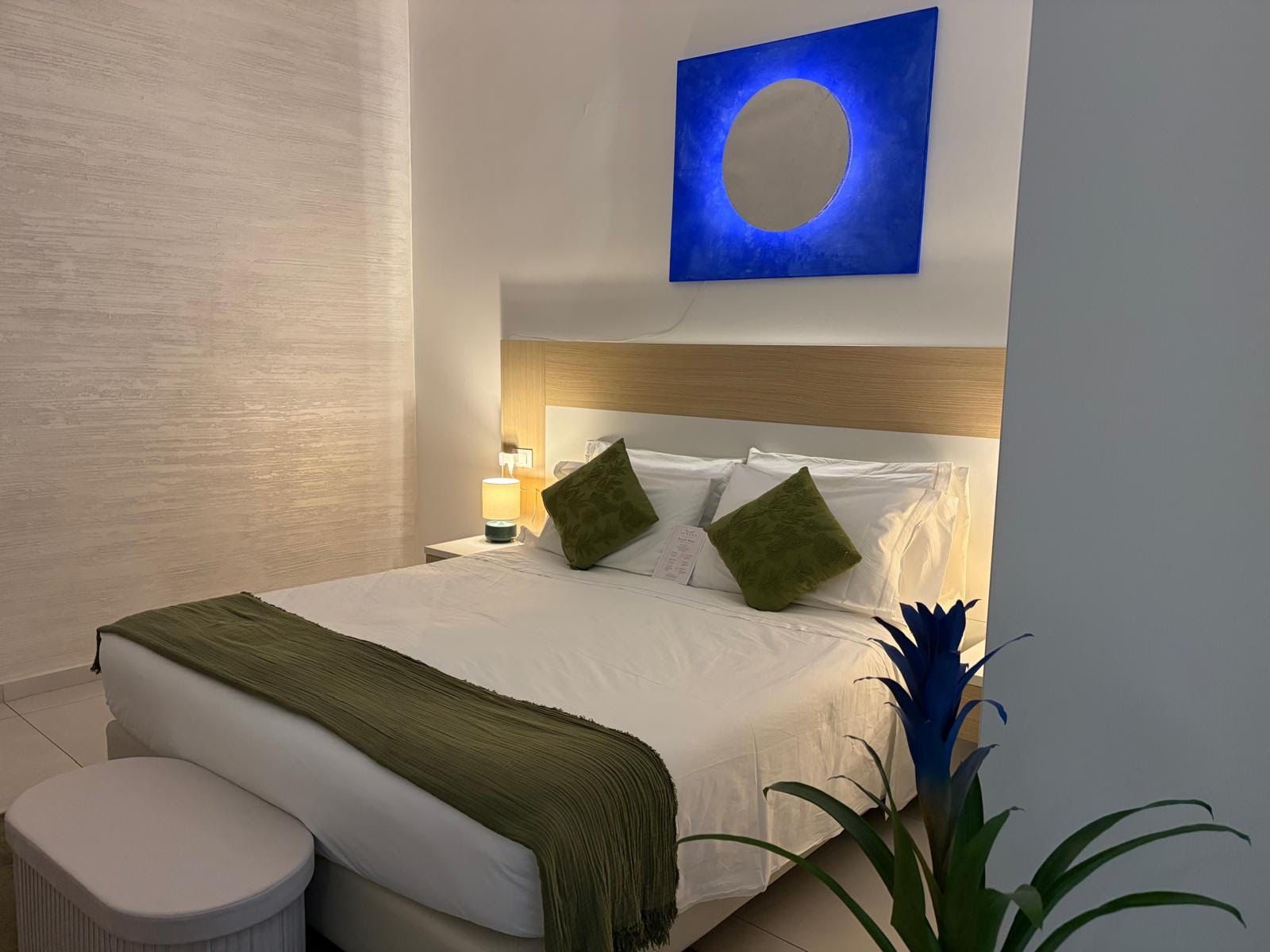 Ein Hotelzimmer mit Bett, Nachttisch, Lampe, einer Zimmerpflanze und einem Wandbild.