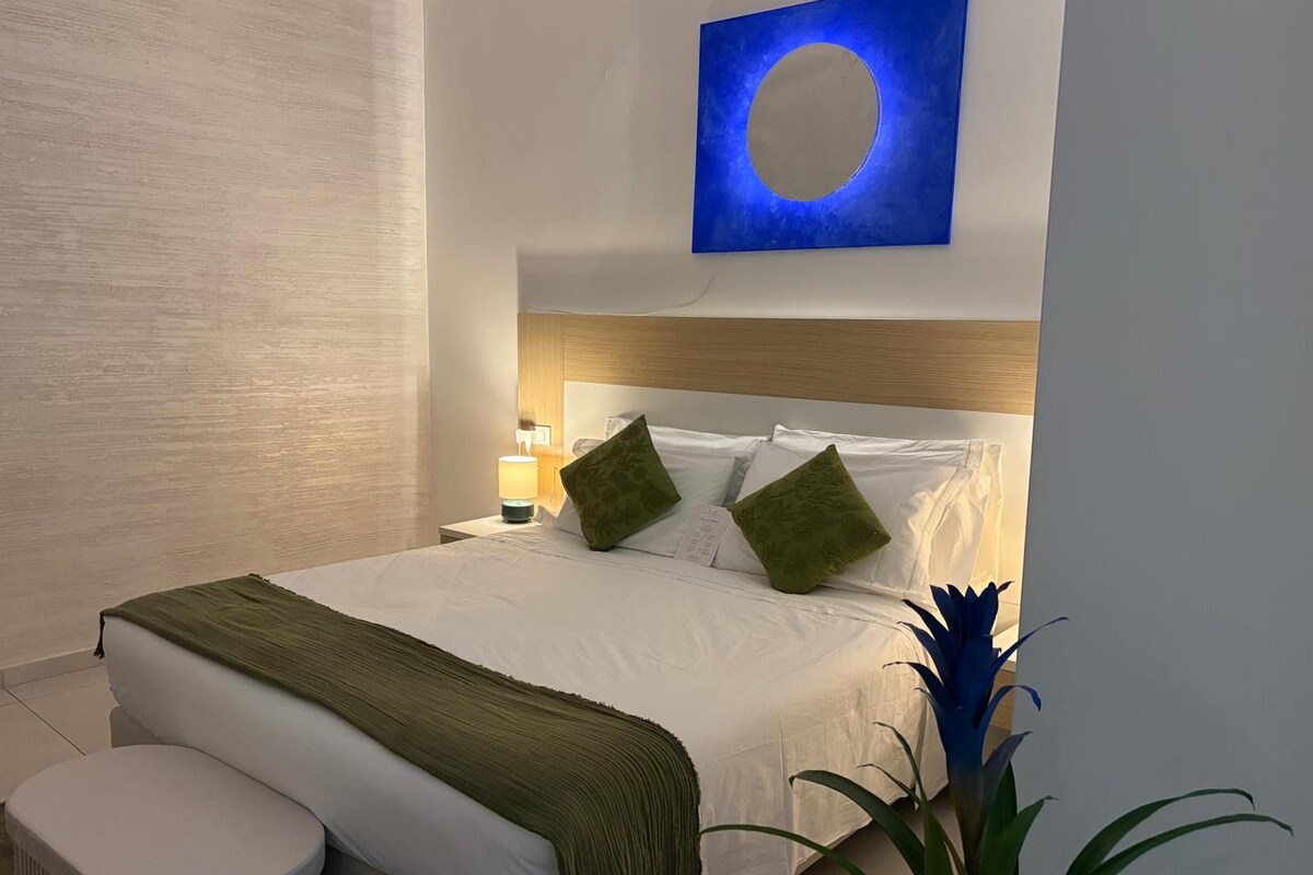 A hotel room with a bed, a nightstand, a lamp, a green plant, and a wall art piece.