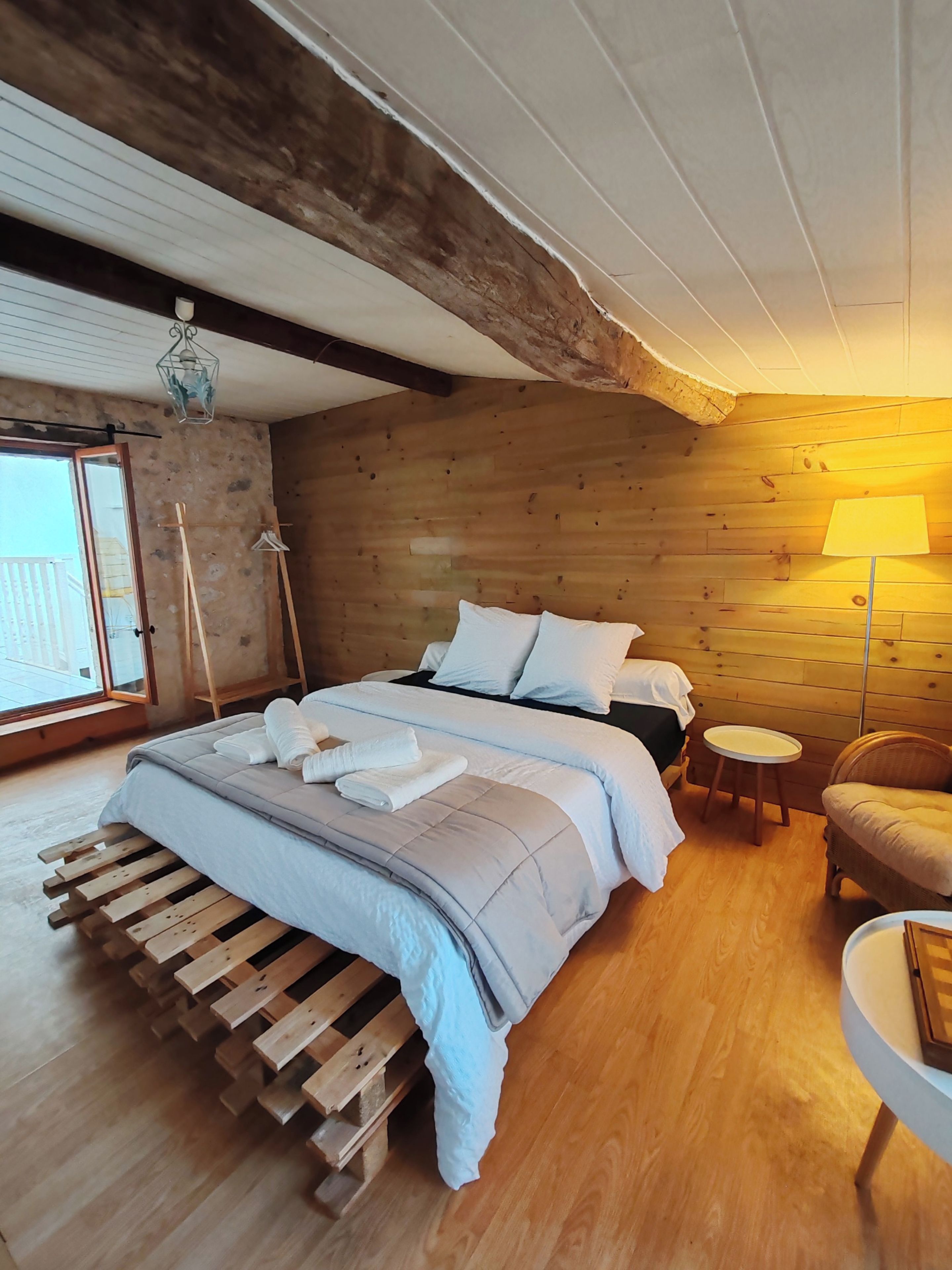 A bedroom with a wooden bed, a bedside table, a chair, and a view through a window.