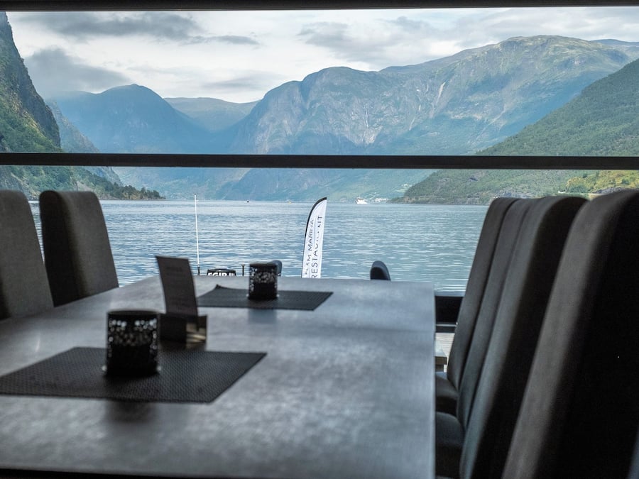 A dining area with a view of a lake and mountains. There are tables with chairs, a flag, and a menu stand.