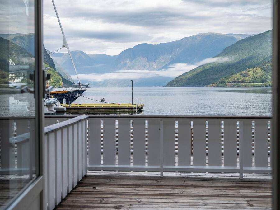 A wooden deck with a railing, overlooking a calm body of water with mountains in the background.
