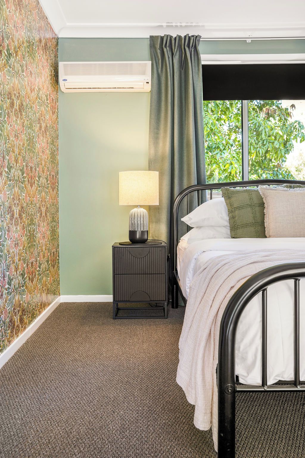 A bedroom with a bed, a bedside table with a lamp, a window with curtains, and a wall with a floral pattern.