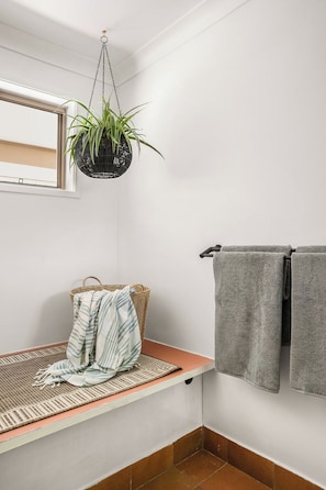 Un baño con una planta colgante, un perchero con dos toallas, una cesta tejida y un banco con una toalla a rayas.