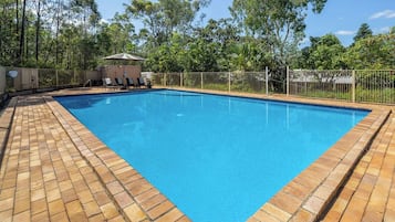 A rectangular swimming pool with a tiled edge, surrounded by a fence, and a covered seating area with chairs and a table.