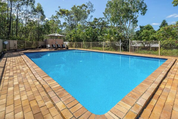 A rectangular swimming pool with a tiled edge, surrounded by a fence, and a covered seating area with chairs and a table.