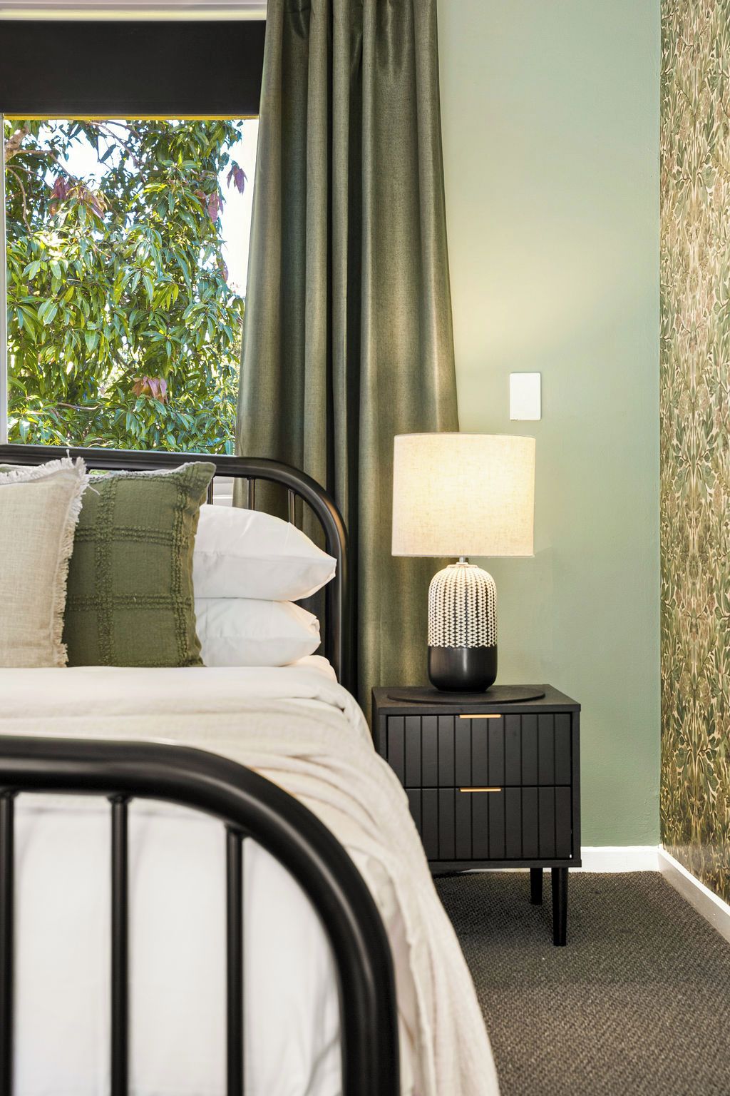 A bedroom with a bed, a nightstand with a lamp, and a view of greenery outside.