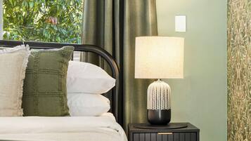 A bedroom with a bed, a nightstand with a lamp, and a view of greenery outside.