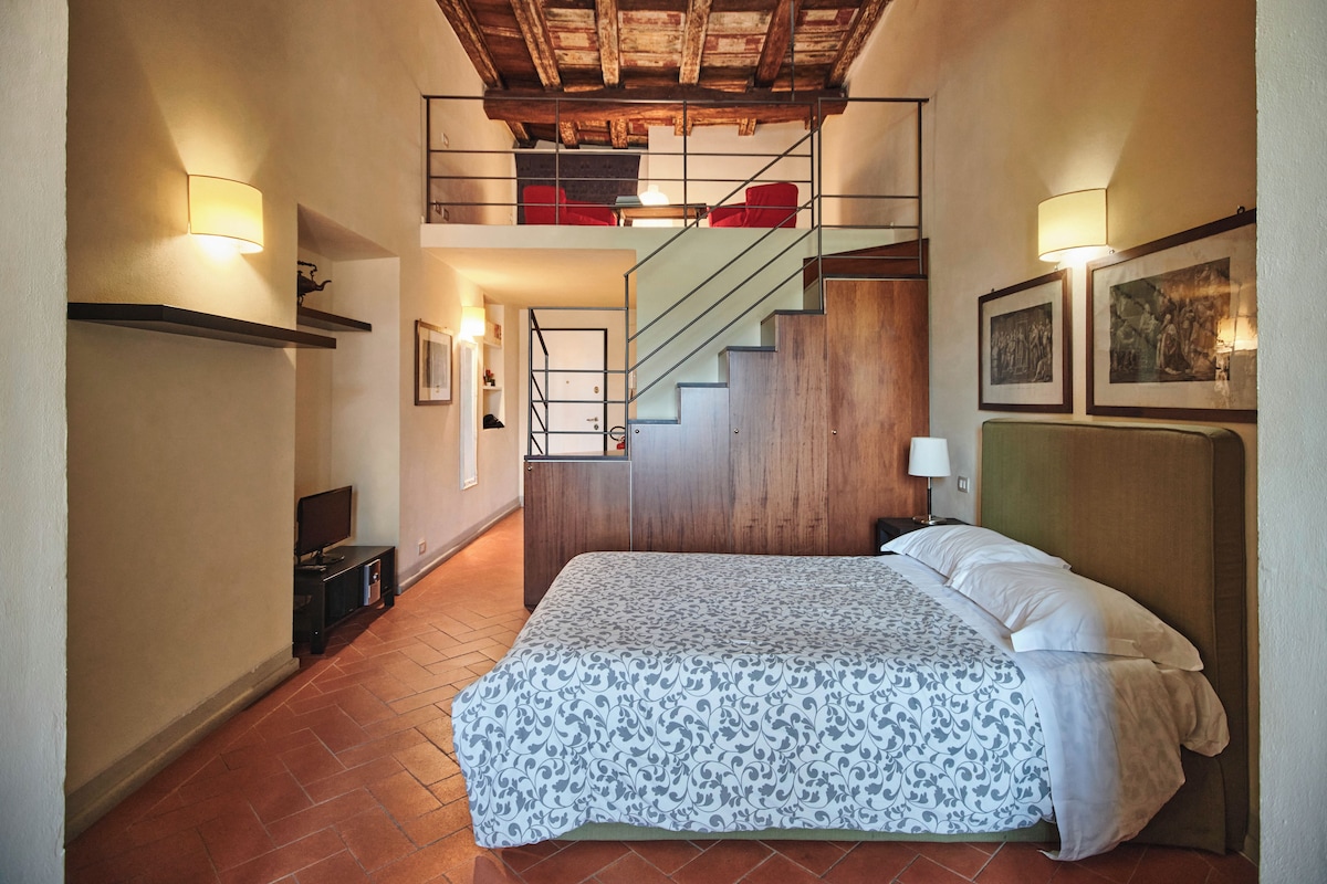 Una habitación de hotel con una cama, un escritorio, una silla, una escalera y una pared decorada con cuadros.