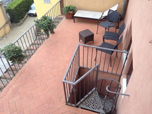 Un balcón con suelo de baldosas, baranda de metal, una mesita, dos sillas y una planta en maceta.