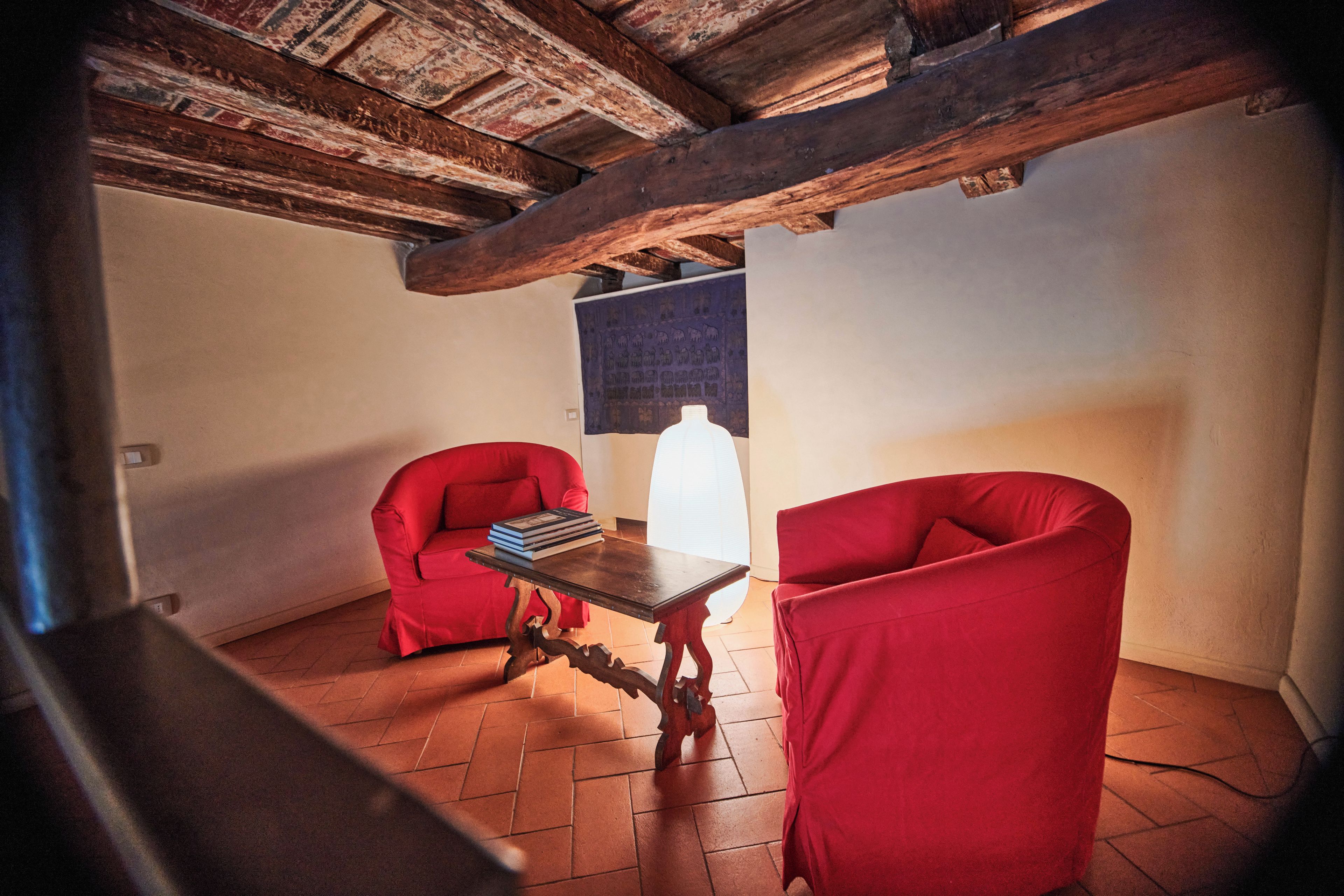 Ein Zimmer mit Holzbalkendecke, zwei roten Stühlen, einem kleinen Tisch mit Büchern und einem Fenster mit Vorhang.